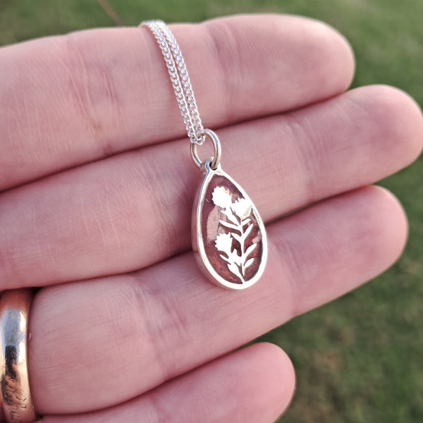 Silver necklace with a teardrop-shaped pendant featuring a floral design, held in a hand against a blurred green background.