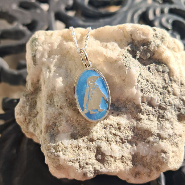 Penguin pendant on a blue background hanging from a chain on a textured stone surface.