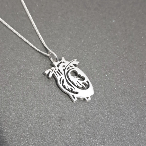 A silver pendant necklace featuring a stylized cutout anatomical heart design, shown on a textured dark gray background.