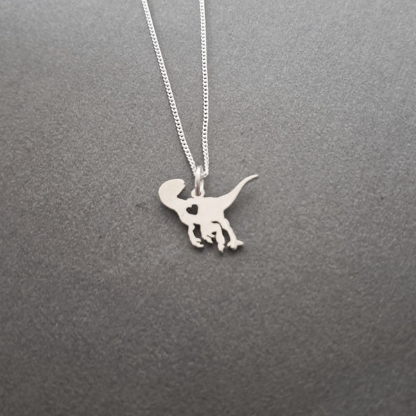 A silver necklace with a dinosaur silhouette pendant, displayed on a textured dark gray background.