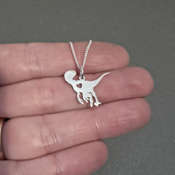 A silver dinosaur pendant necklace with a heart-shaped cutout on a chain, held in a person's hand against a gray background.