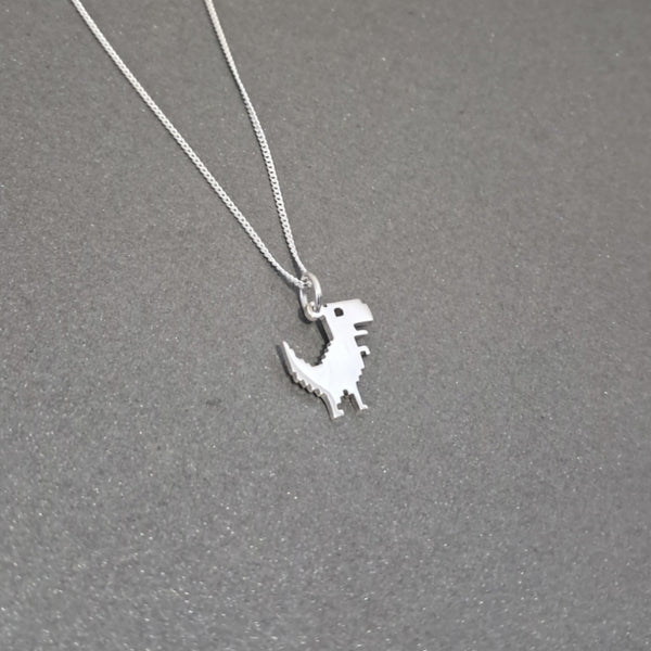 A silver dinosaur pendant necklace on a delicate silver chain, displayed on a textured dark gray background.