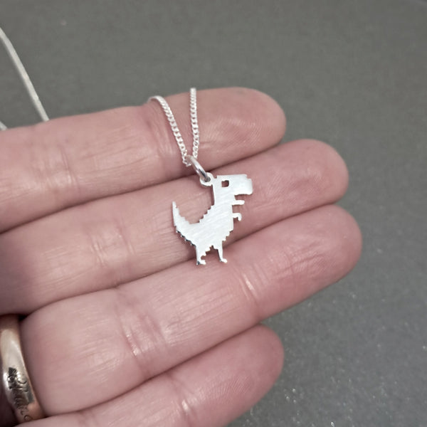 A silver dinosaur pendant necklace held on a person's fingers against a dark gray background.