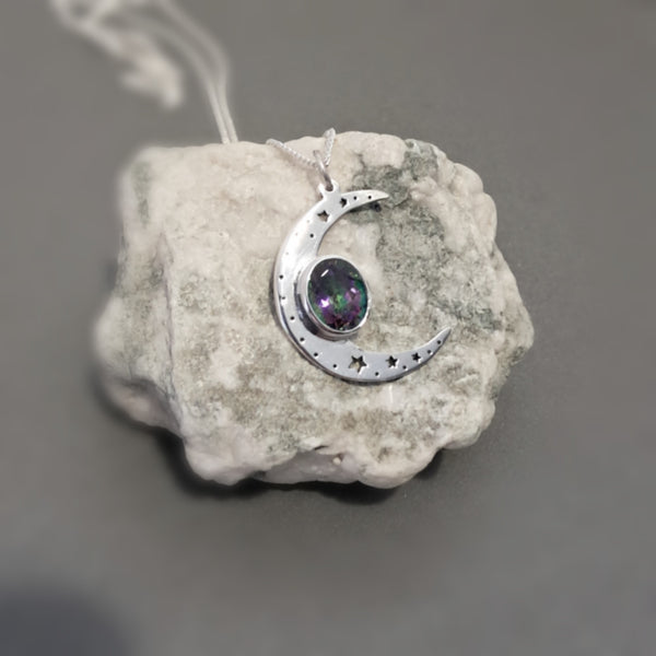 A silver crescent moon pendant with small star cutouts and a round iridescent gemstone, displayed on a textured white stone against a dark gray background.