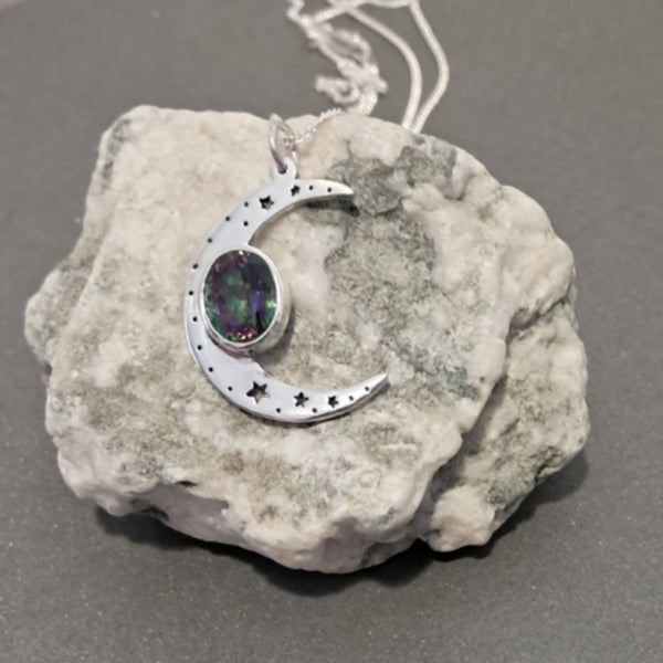 A silver crescent moon pendant necklace with star cutouts and a round iridescent gemstone, displayed on a textured white rock against a dark gray background.