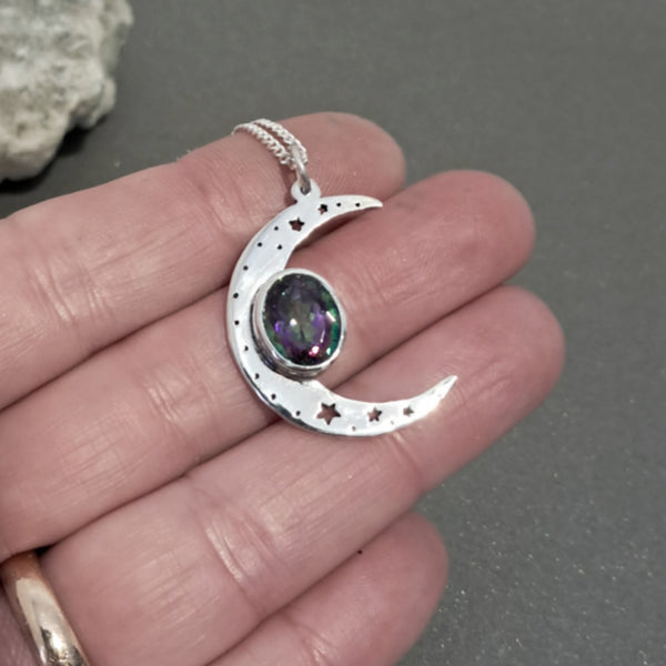 A silver crescent moon pendant with star cutouts and an oval iridescent gemstone, held in a person's hand.