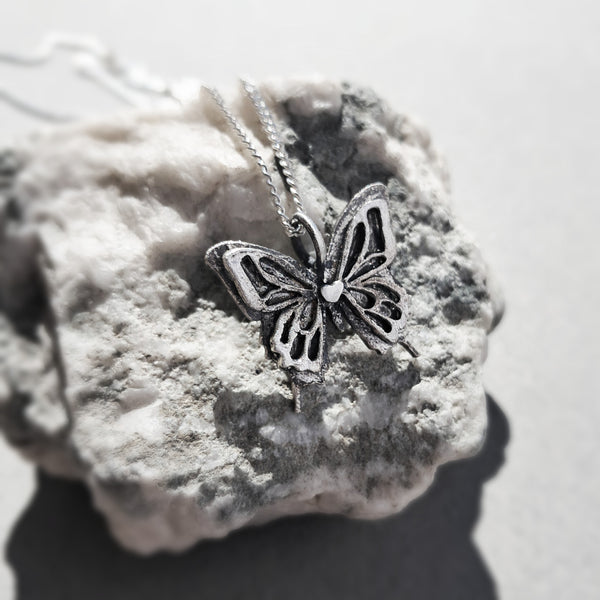 A silver butterfly pendant necklace with an oxidized finish and a small  heart detail, resting on a textured white stone against a light gray background.
