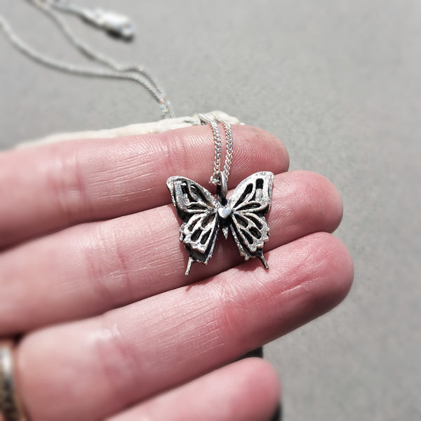 A close-up of a hand holding a silver butterfly pendant necklace with an oxidized finish, set against a neutral gray background.
