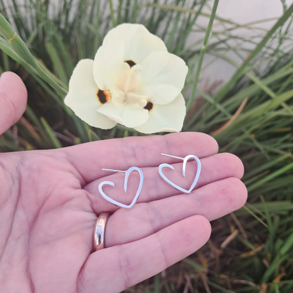 A person's hand holding a pair of small silver heart-shaped hoop earrings, with a pale yellow flower and green grass blurred in the background.